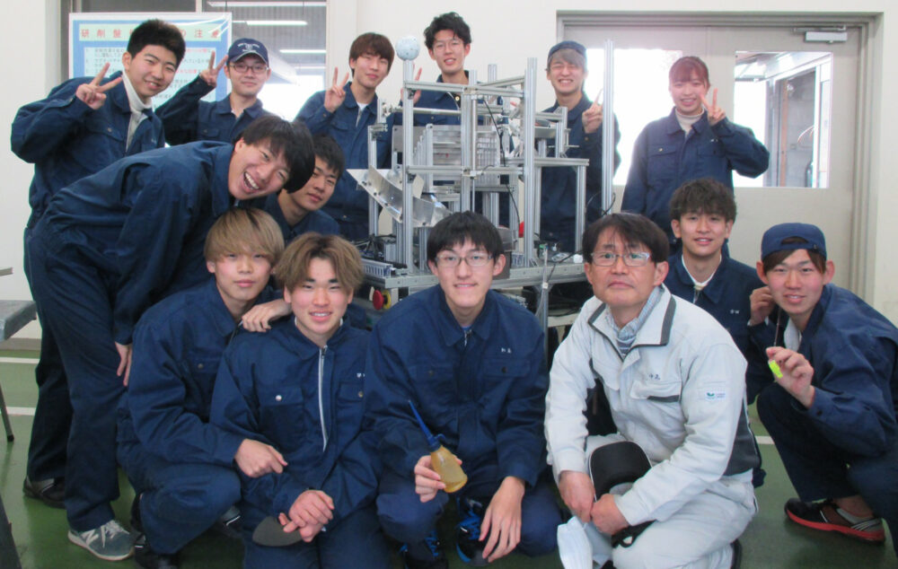 紺色の作業着を着た15名程の学生たちが中央にあるロボットを囲んだ集合写真